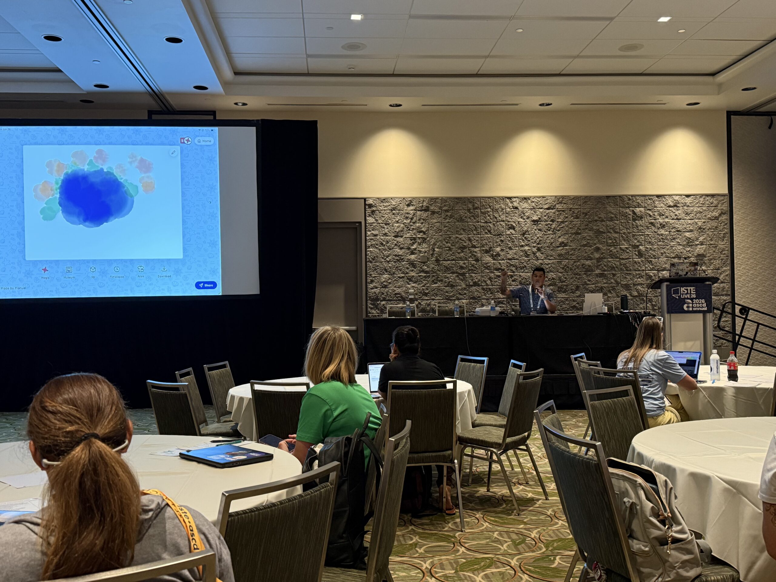 Conference talk with presenter gesturing beside a projected color graphic, seated attendees watching at round tables.