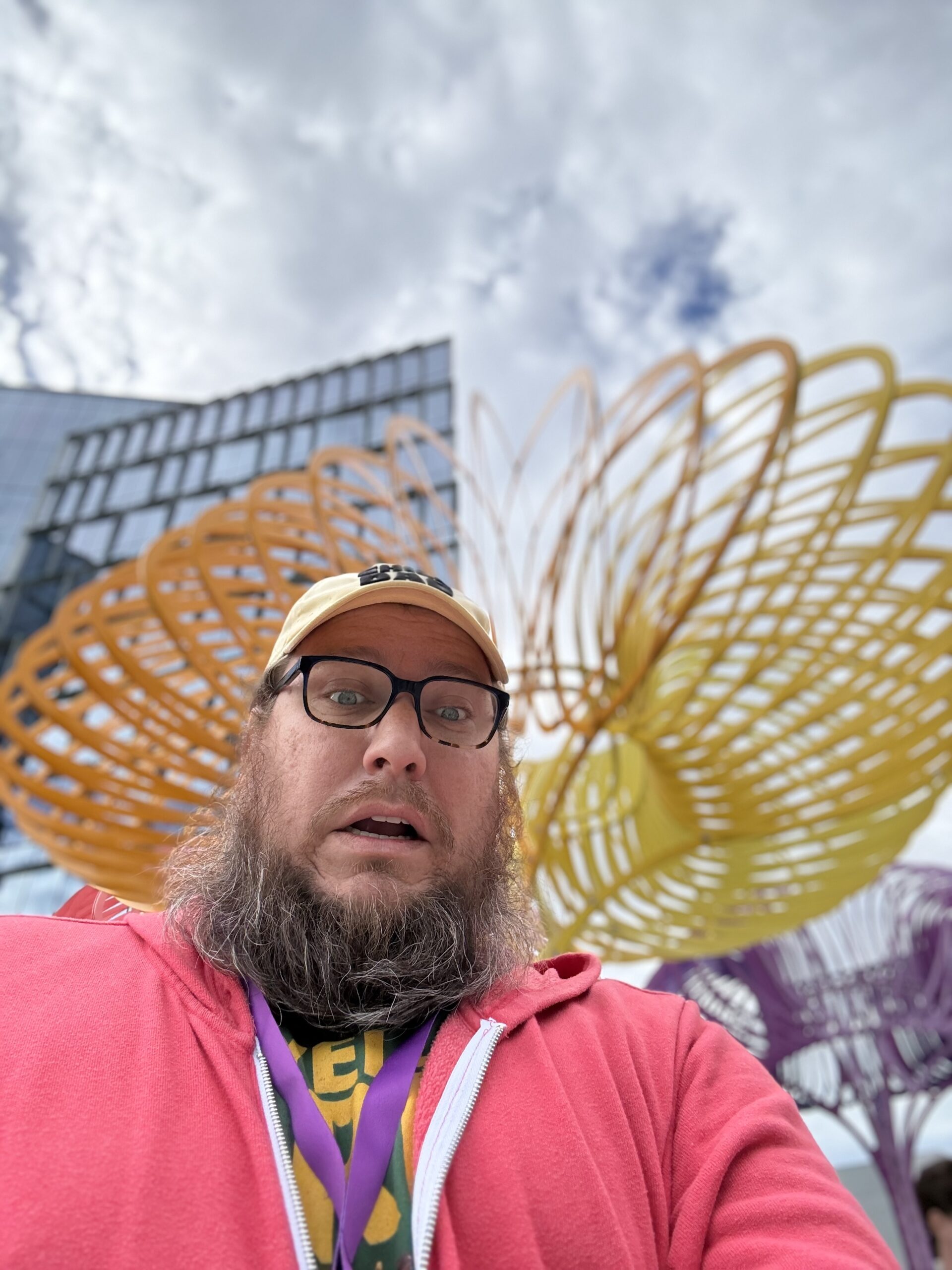Selfie of a bearded man in a pink hoodie and cap, looking surprised, with colorful spiral sculpture and glass building under cloudy sky.