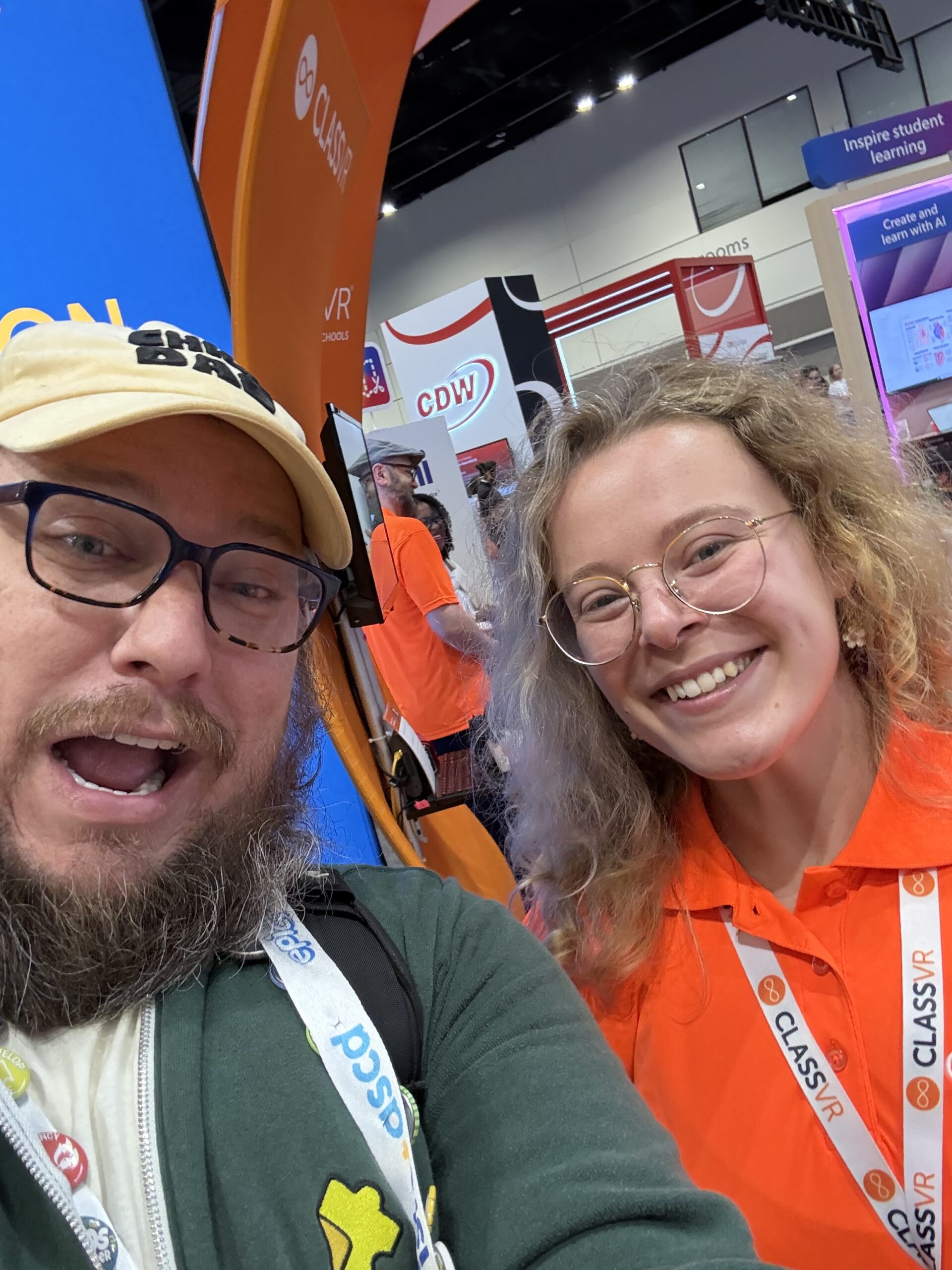 Two smiling attendees take a selfie at a ClassVR booth at a tech expo, with colorful signs and displays behind them.