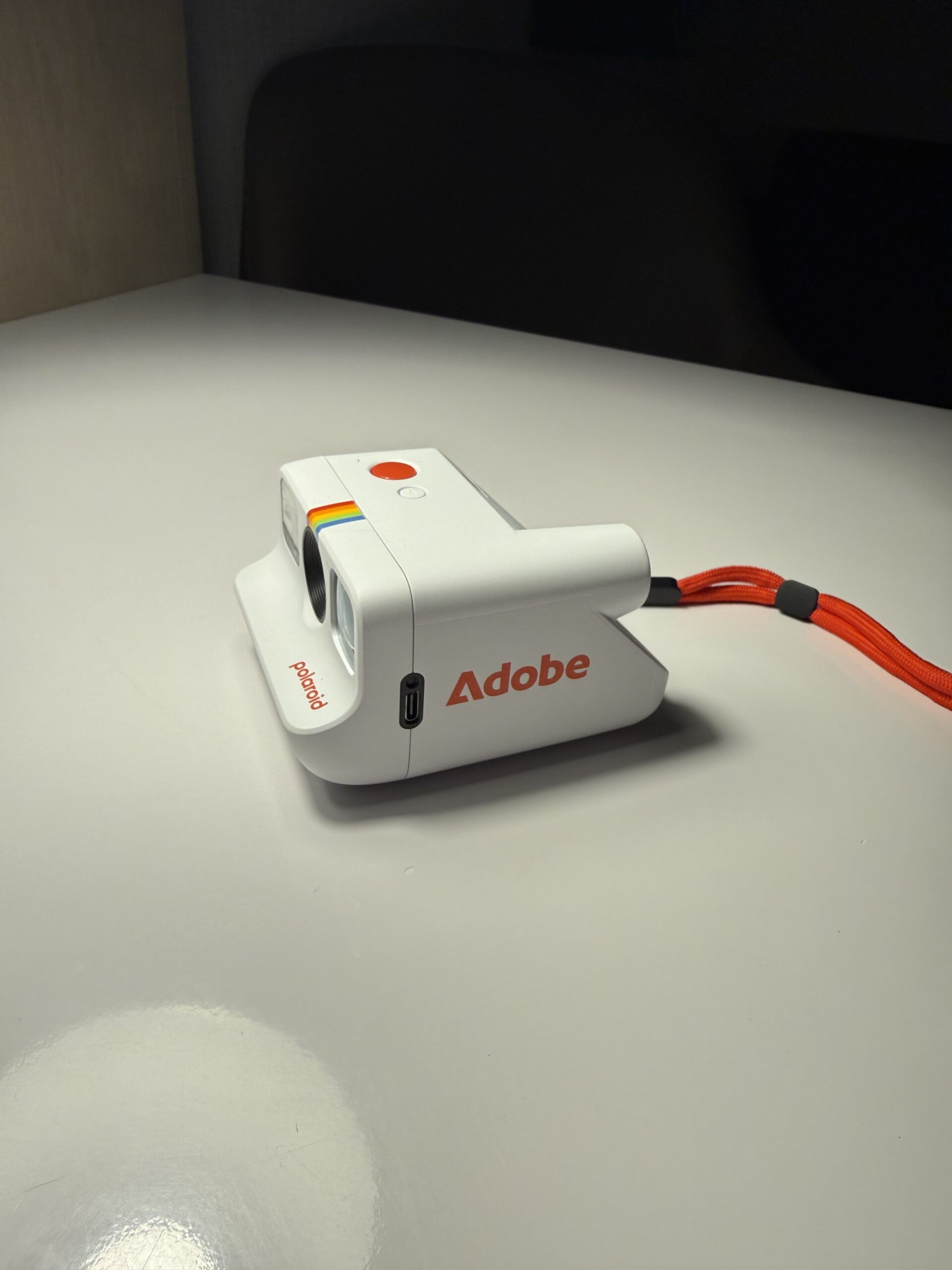 White Polaroid camera with Adobe logo and orange wrist strap on a table, in a dim minimalist room.