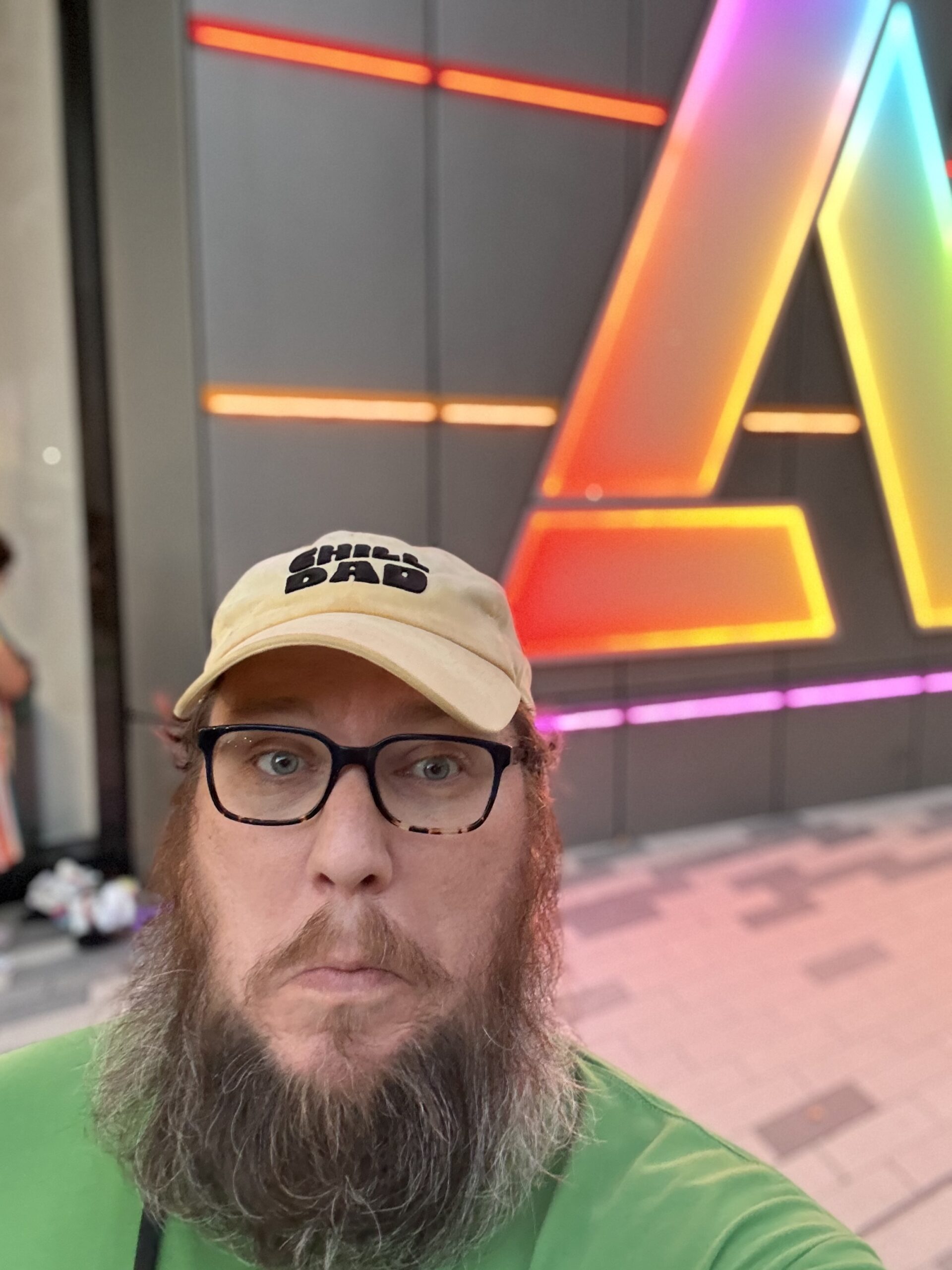 Bearded man in glasses and green shirt takes a selfie in front of a neon A sign, wearing a cap that says CHILL DAD.