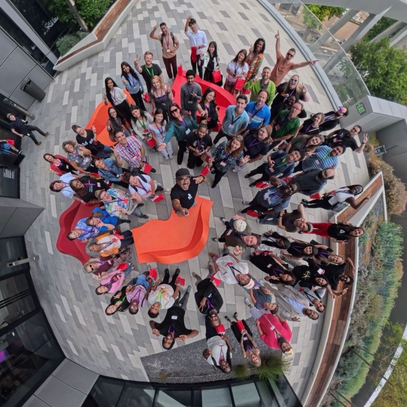 Overhead view of a smiling group waving on a tiled patio around bright orange and red chairs, like a team gathering.