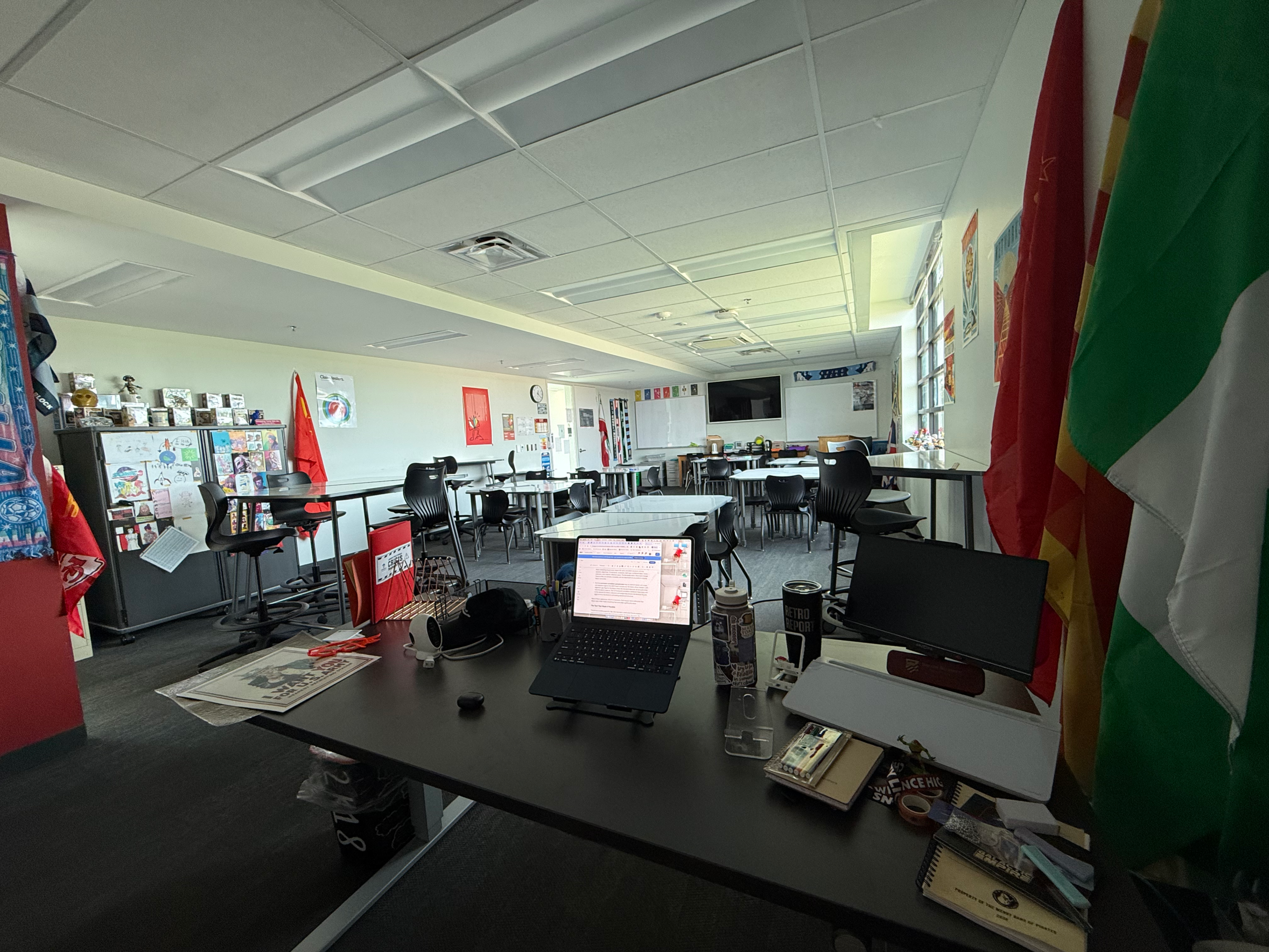 Empty classroom office with laptop and desk in foreground, black chairs, colorful flags, posters, and bright windows.