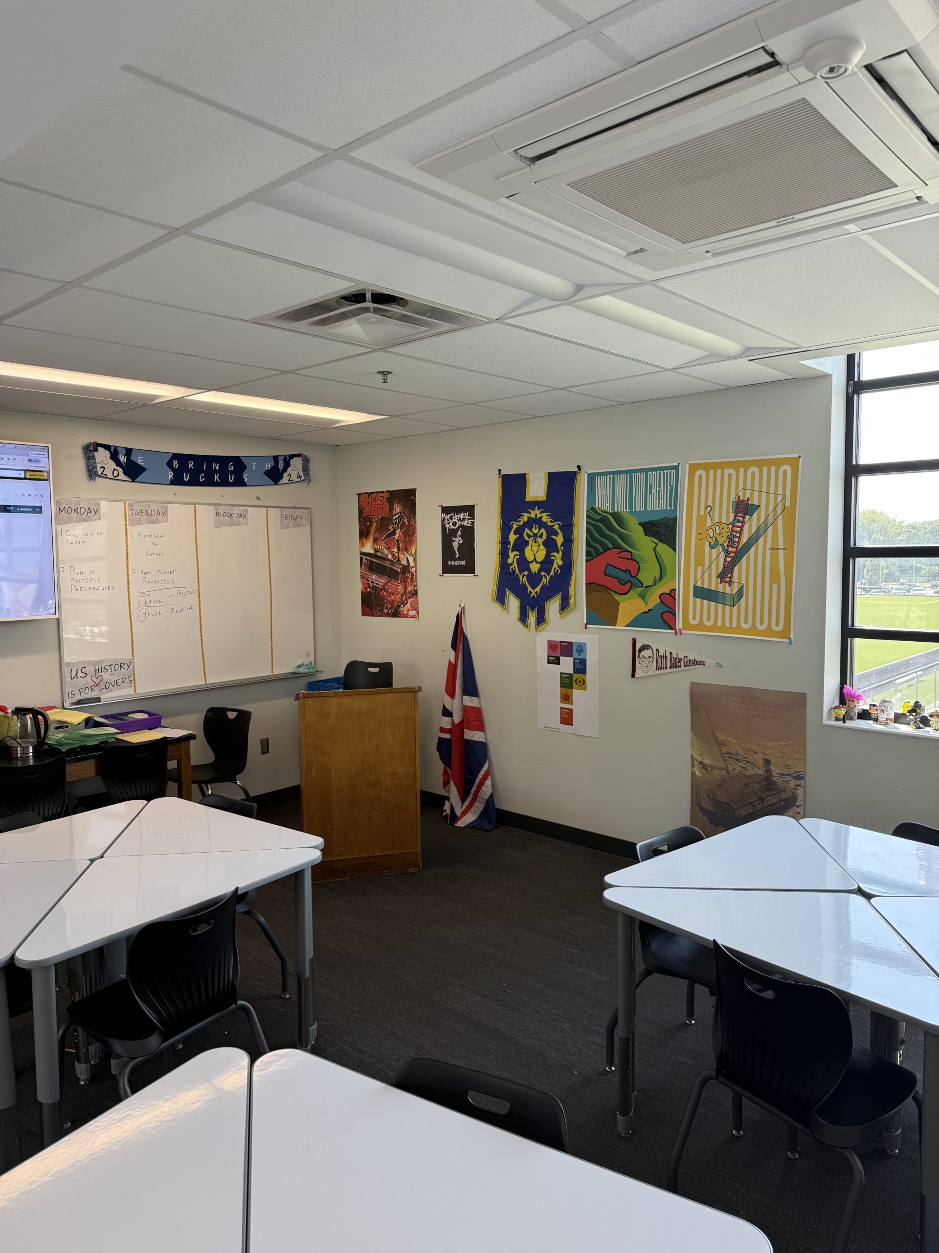 Empty classroom with white tables, black chairs, whiteboard, podium, and colorful posters; sign reads We Bring the Ruckus.