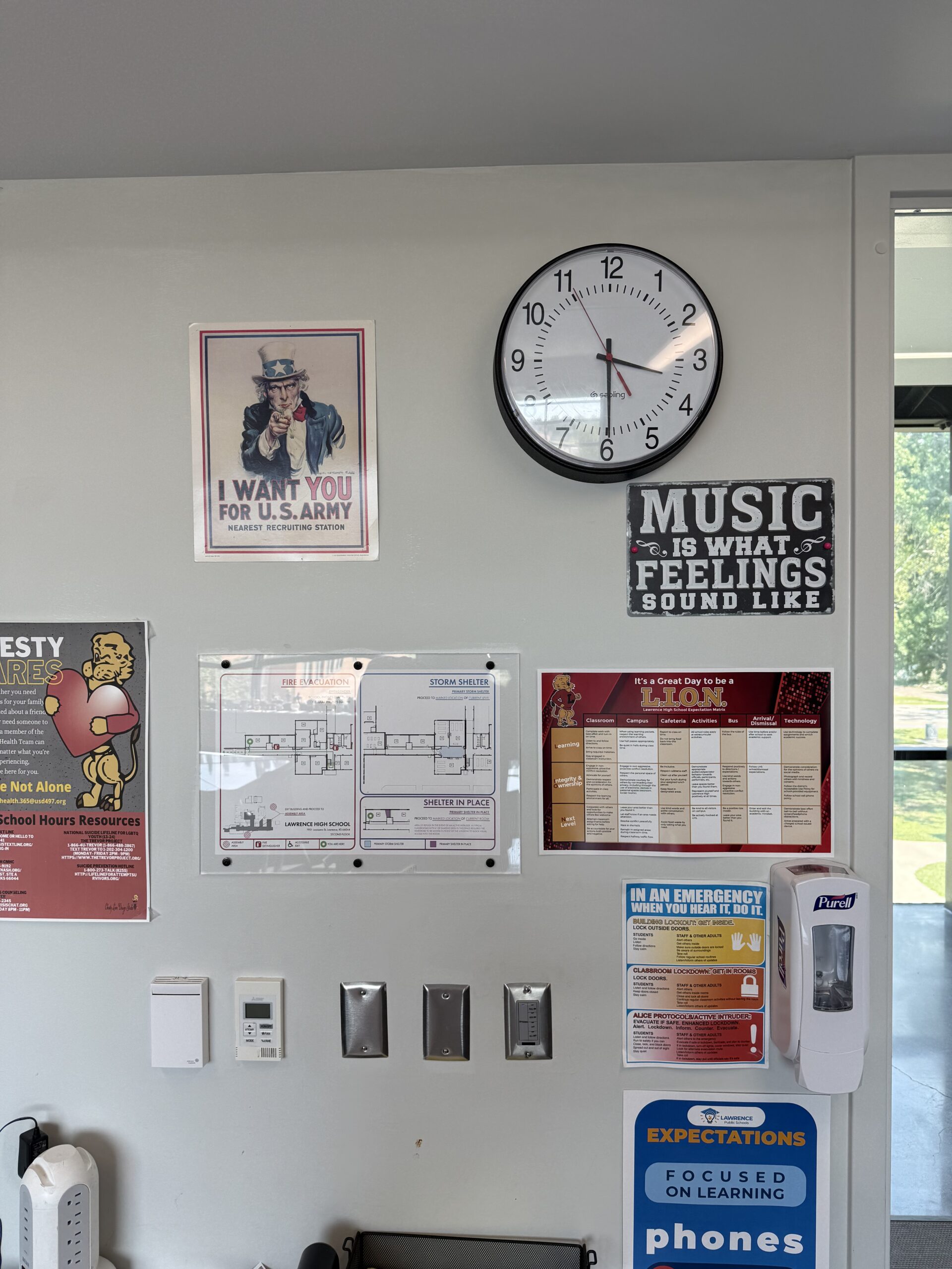 School wall with analog clock, U.S. Army poster, music quote sign, safety maps, emergency info, and hand sanitizer dispenser.